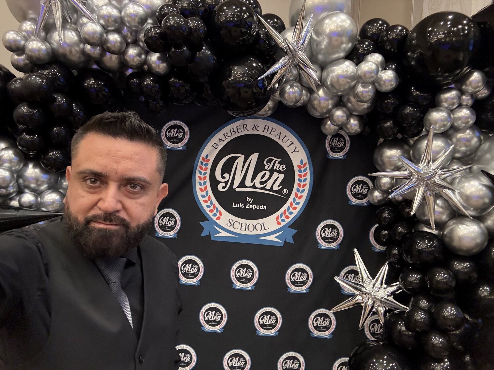 Retrato de Luis Zepeda ante el fondo de The Men Barber School con globos negros y plateados